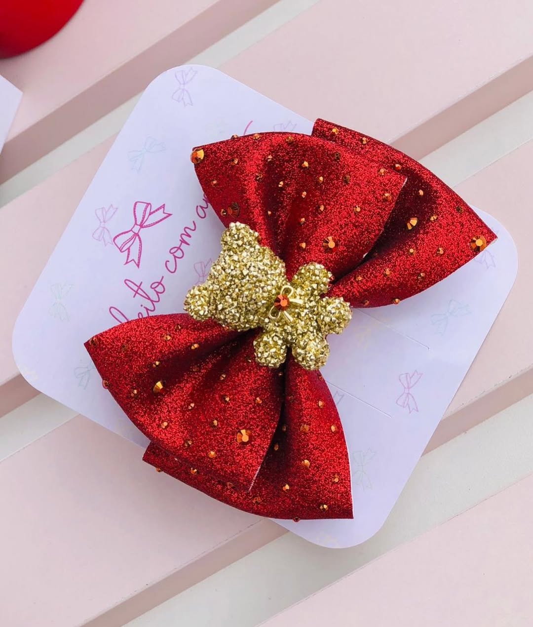 Laço vermelho com glitter e ursinho dourado, feito à mão, sobre cartela de apresentação